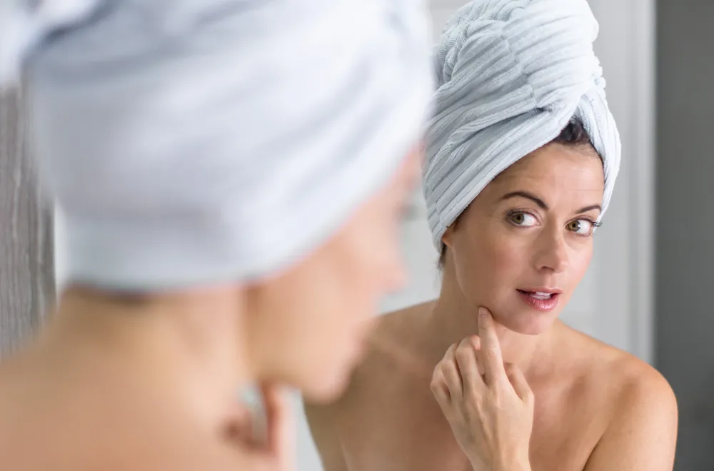 A woman with a towel wrapped around her head is examining her skin in the mirror