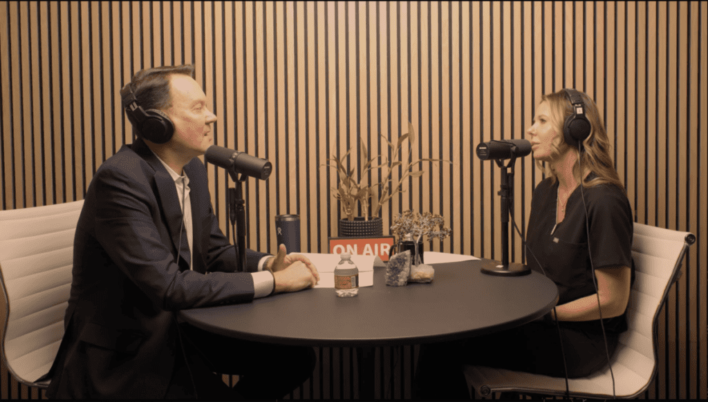 Two people wearing headphones at a podcast table with microphones and a sign that reads "ON AIR."