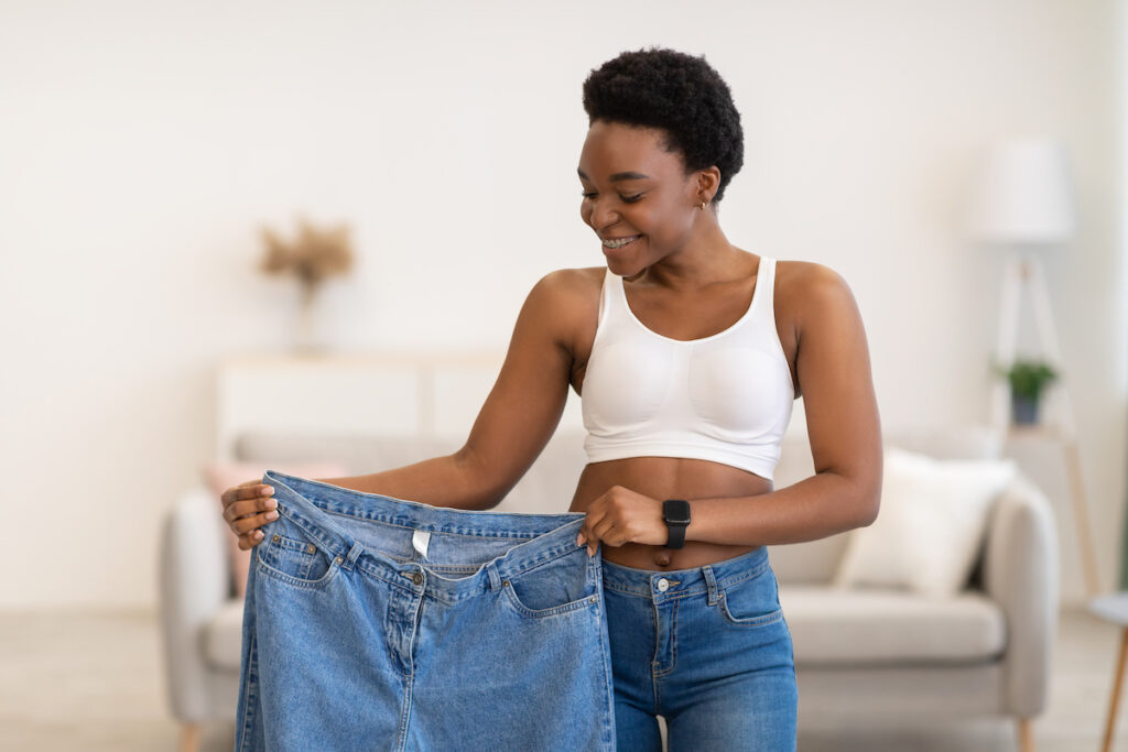 A woman is smiling and holding up a pair of jeans that are too big now that she has lost weight with the help of Xanadu weight loss clinic in Fort Collins